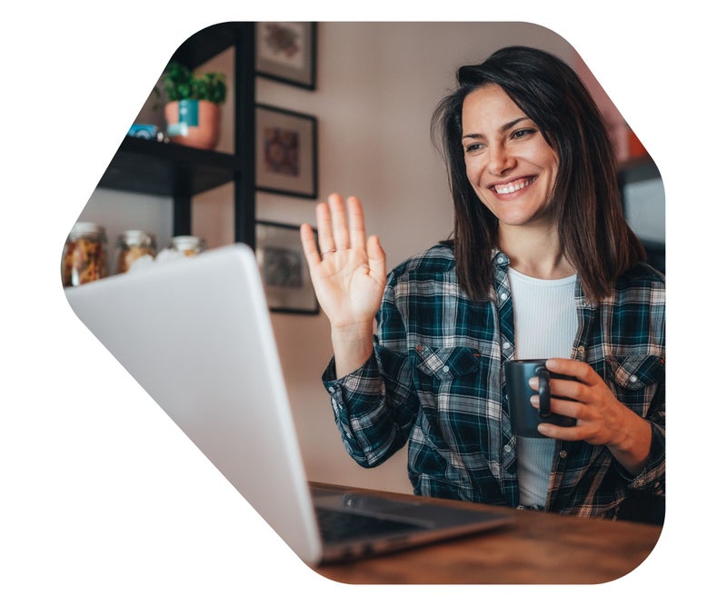 Woman attending an online telehealth psychology session from home, holding a mug and waving at her laptop in a comfortable setting.
