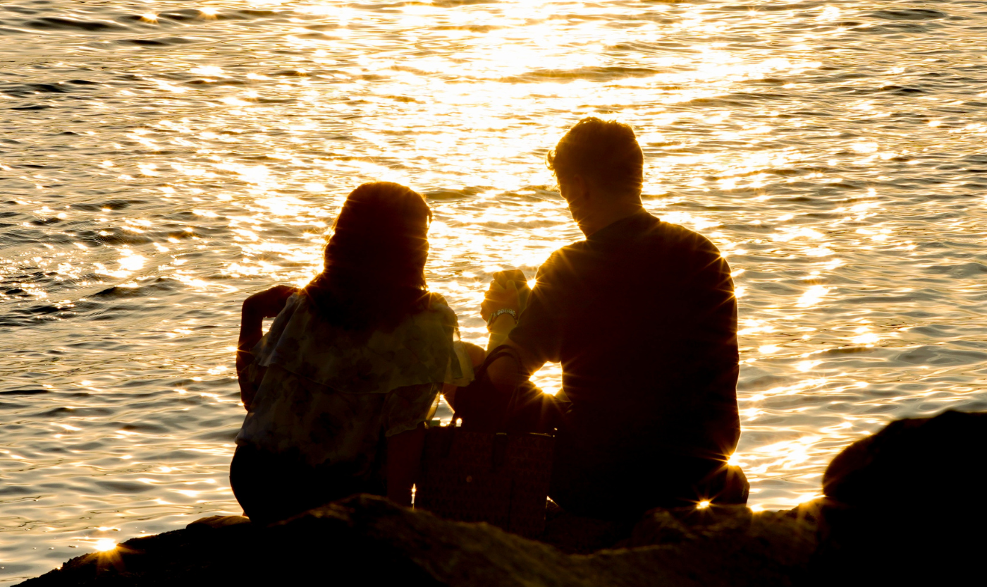 A couple sits in silhouette by the water at sunset, sharing a quiet moment. This scene conveys supportive connection during hard times.