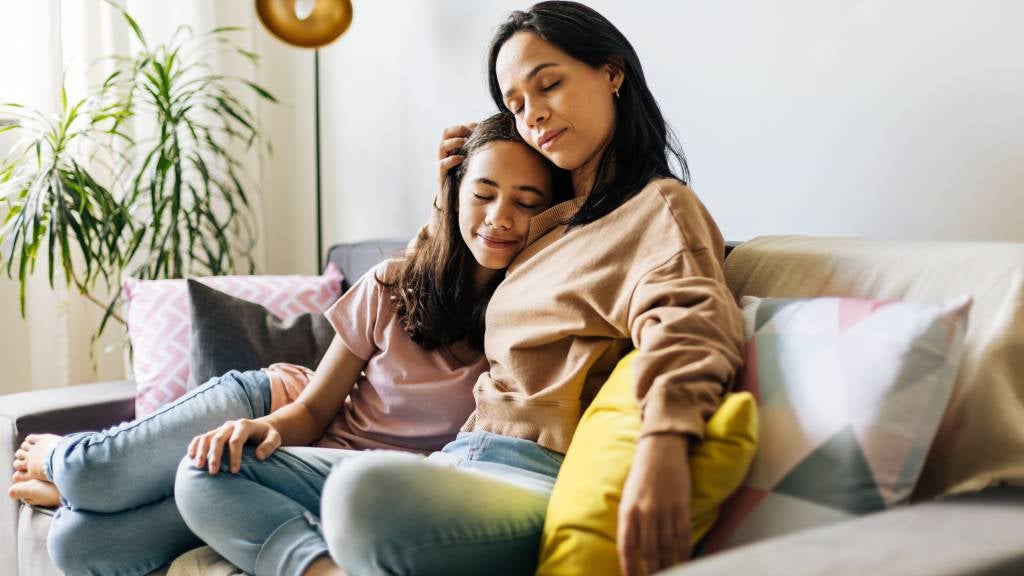 A woman and her daughter with closed eyes sit on their sofa leaning on each other
