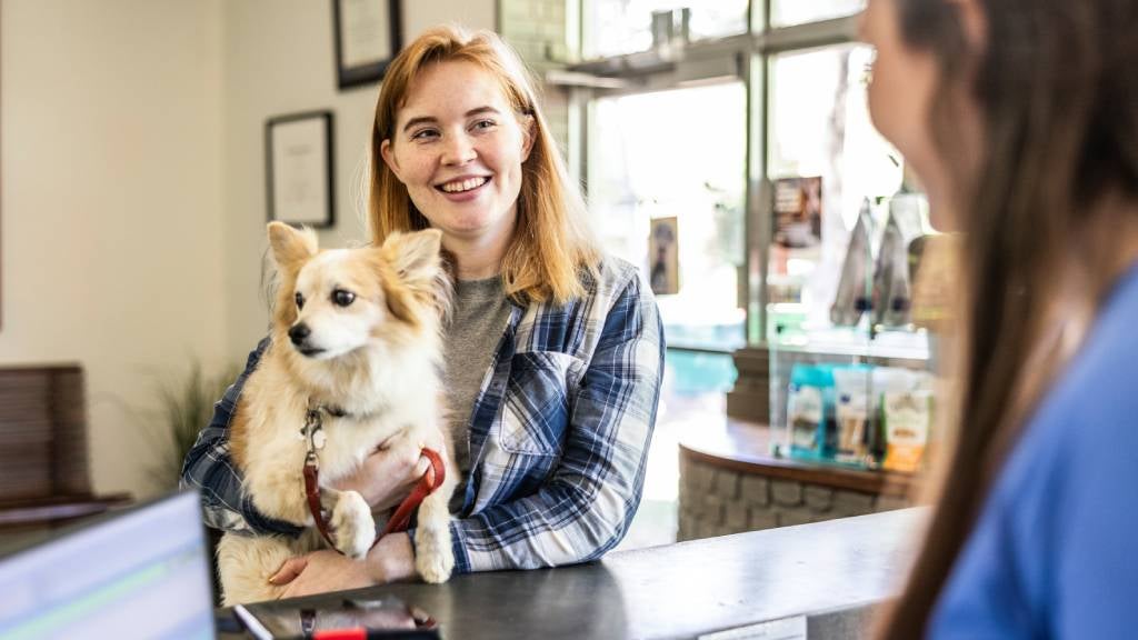A smiling woman stands at a vet clinic reception while holding her small dog 