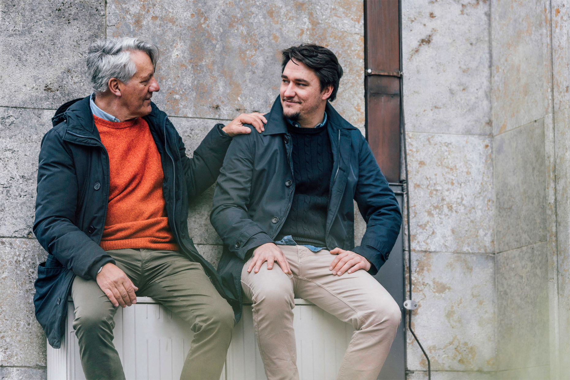 Two men are sitting next to each other. The older man looking at younger man. The younger man is looking at the older man and smiling. The older man has his hand on the shoulder of the younger man.