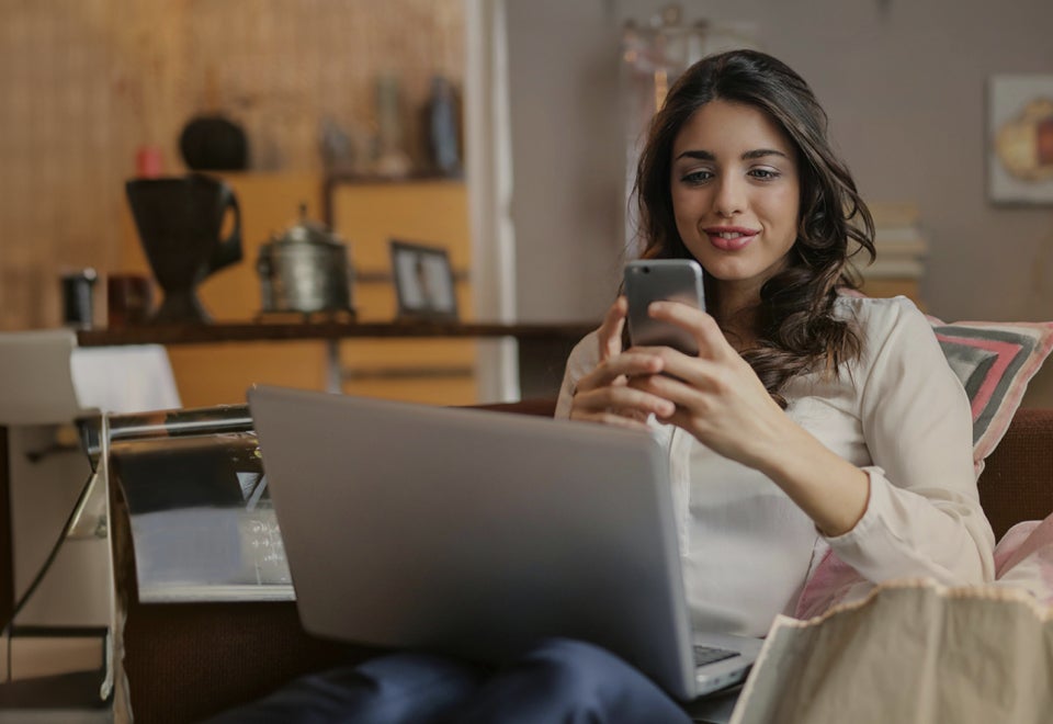 Regional Australia Bank | A smiling woman sitting in a chair with a laptop on her lap while she is using a smartphone | Devotion