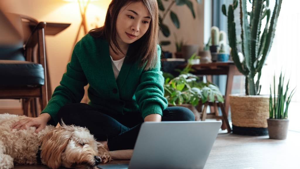 A woman sits on the floor and pets her dog while typing on her laptop