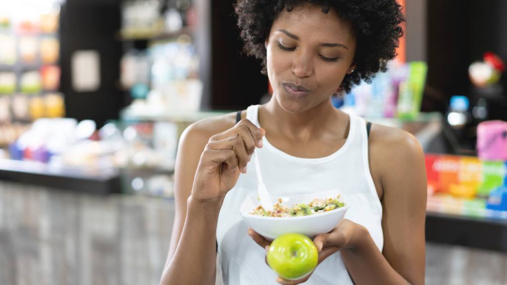 Woman in white singlet eating bowl of muesli holding a green apple