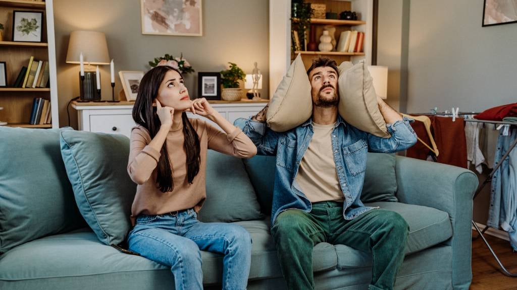 Young couple sit on couch, covering their ears while staring at the roof, hearing a noisy neighbour upstairs