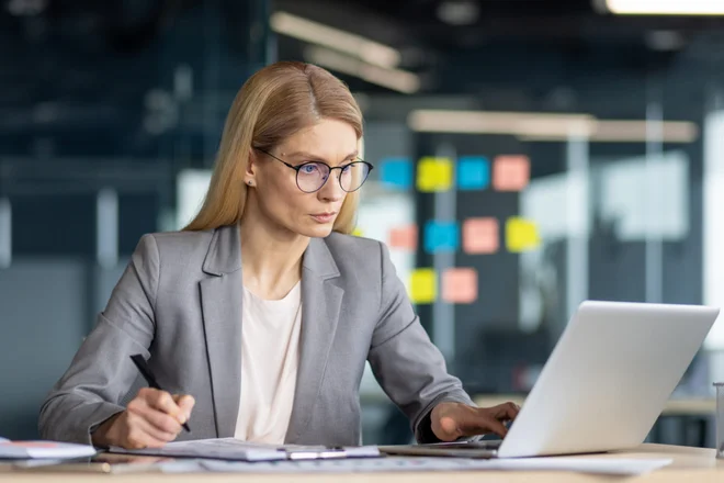 A professional businesswoman engages in serious work, meticulously analyzing paperwork and thinking at her office desk with a laptop.