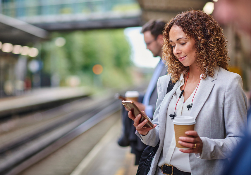 Women reading an email on her phone while at the train station, holding a coffee in her other hand.