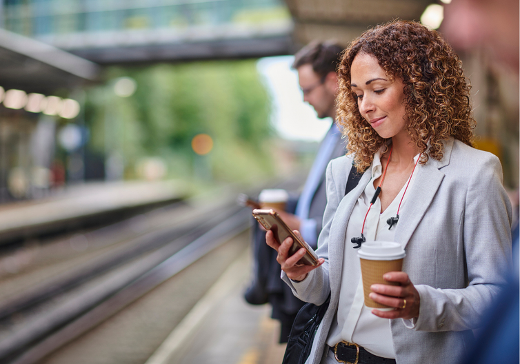 Women reading an email on her phone while at the train station, holding a coffee in her other hand.