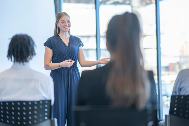 A young female business professional stands at the front of a conference room as she makes a presentation to her colleagues. She is dressed professionally and is using her hands to help articulate her points.