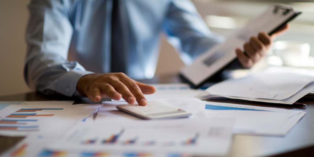 Man in business clothing types into calculator, holding a clipboard, surrounded by sheets of paper.