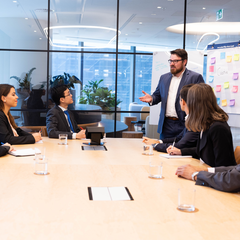 Meeting with six business people sitting around a board table, with whiteboard. A man standing in grey suit with arms open and talking