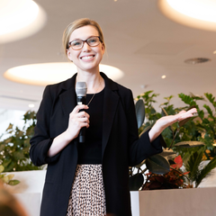 Business woman standing with microphone and outstretched hand, and with hair pulled up and with glasses, in dress and jacket