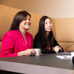 Two business women sitting in an office both looking to another person. One women is a bright pink jacket smiling and another women in black looking away intently