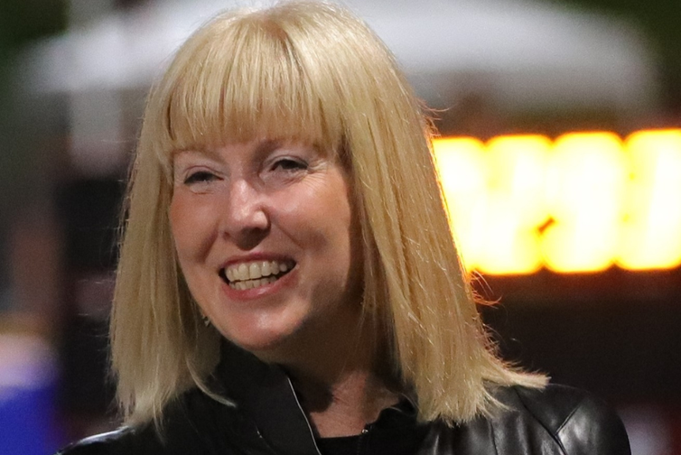 A photo of Jan Swinhoe looking to left and smiling. Photo shoulders up wearing black jacket on and blonde hair to shoulders