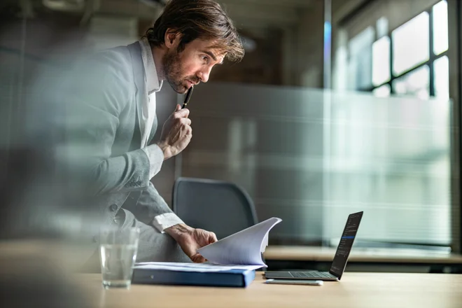 Male worker comparing documents with computer data while sitting on a desk in the office.