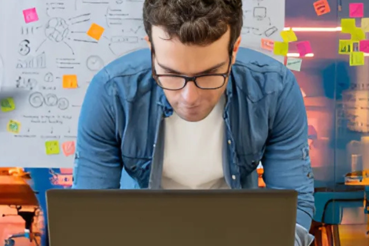 man leans over laptop with whiteboard behind him