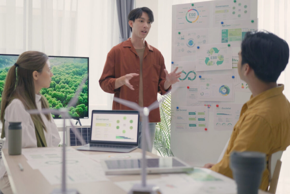 a man stands talking to a man and woman, surrounded by models of wind generators and print outs of charts.
