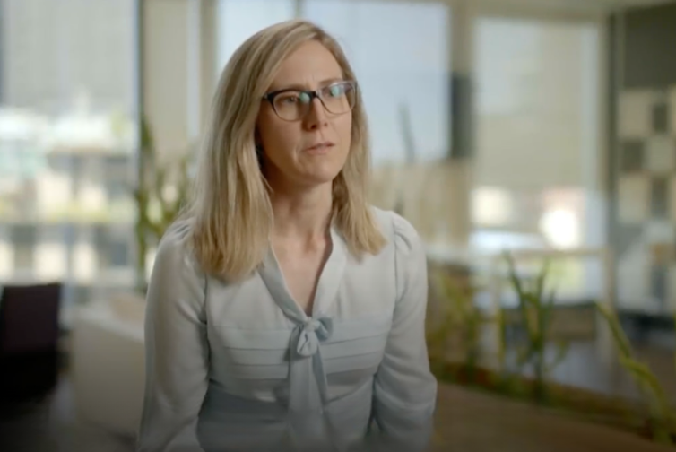 Femalw with blonde haor to shoulders and white shirt with bow, talking.
