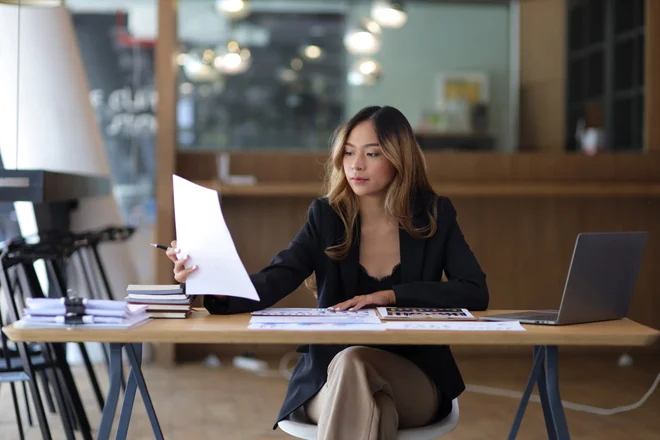 Smiling Asian businesswoman working on laptop and reading documents on desk in office.
