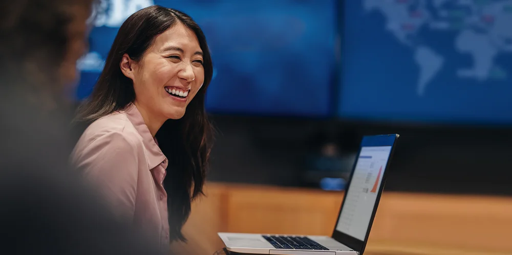 Lady with black hair smiling infront of globe map with laptop open in front of her.