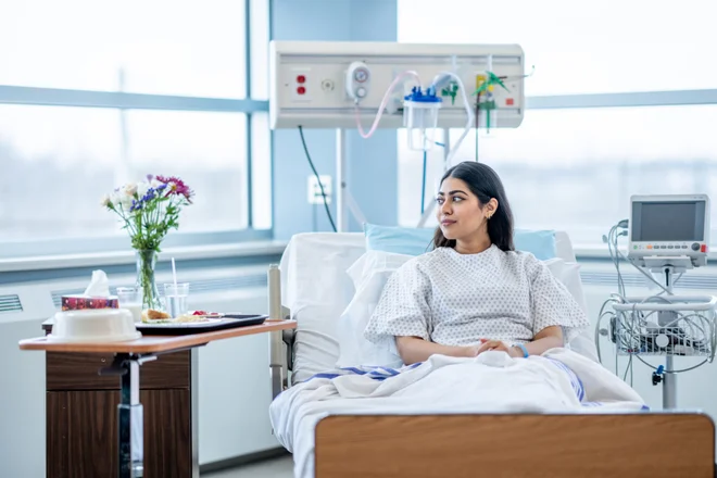 A young woman of Middle Eastern decent sits up in her hospital bed after surgery.  She is wearing a gown and is tucked warmly under a blanket and she looks down contemplatively.