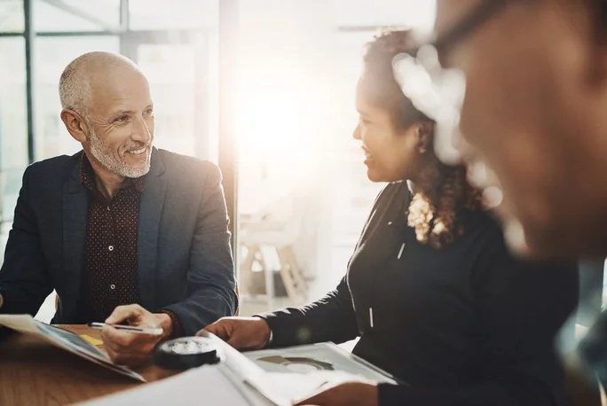 Man and woman meeting at conference table. Light streaming in window behind.