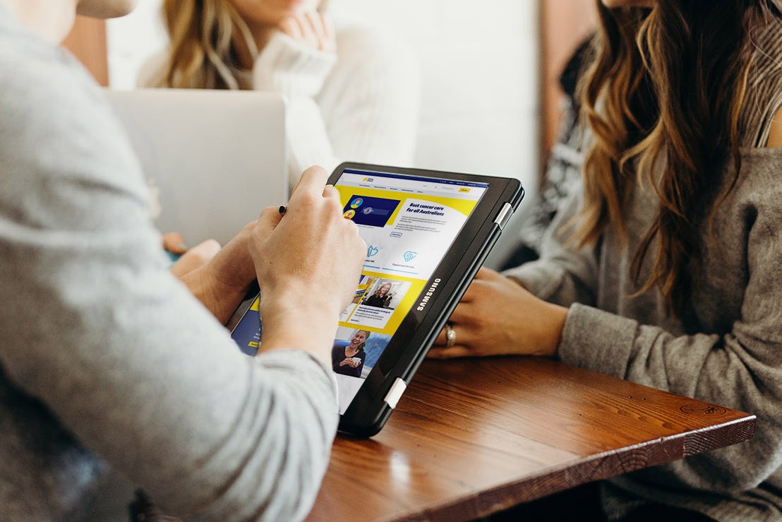 Three people smiling, one is holding an iPad showing a Cancer Council page and another has a laptop open.