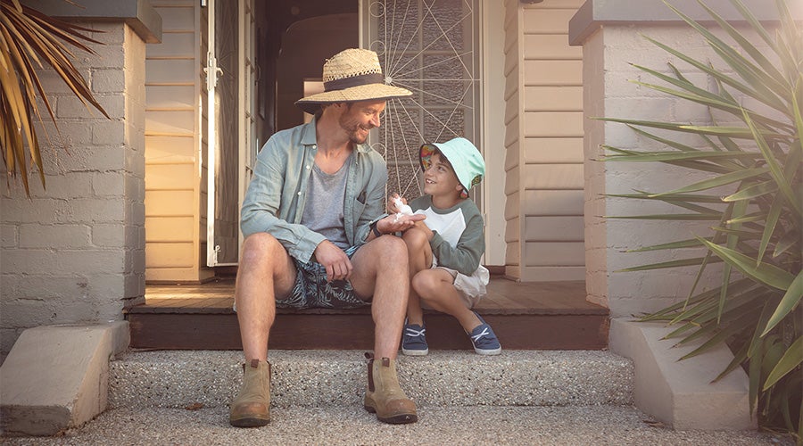 A man and a child sitting side by side on a porch step. They are both wearing long sleeve tops and wide-brimmed hats. The boy is holding a bottle of sunscreen and squirting some onto the man's palm. 