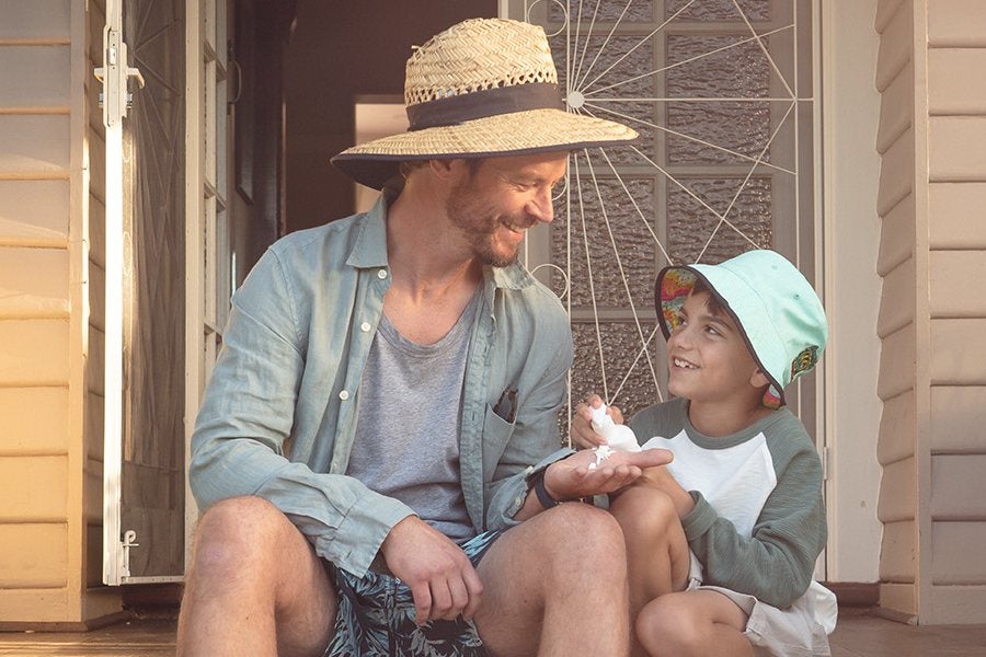 A man and a child sitting side by side on a porch step. They are both wearing long sleeve tops and wide-brimmed hats. The boy is holding a bottle of sunscreen and squirting some onto the man's palm. 