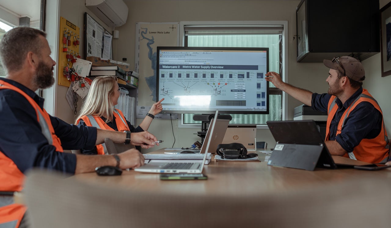 Watercare hydrologists and dam technicians conduct a weekly catch up at their Lower Huia dam office, discussing flow rates and water supply levels displayed on screens.