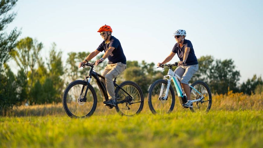 Seniors on two electric bikes, in a natural park setting.