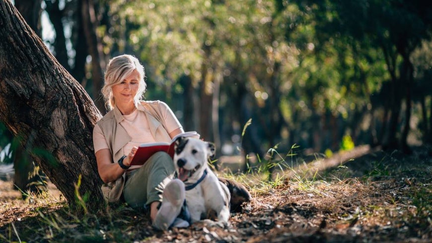 Older Australian woman sits under tree reading a book, in nature with her dog