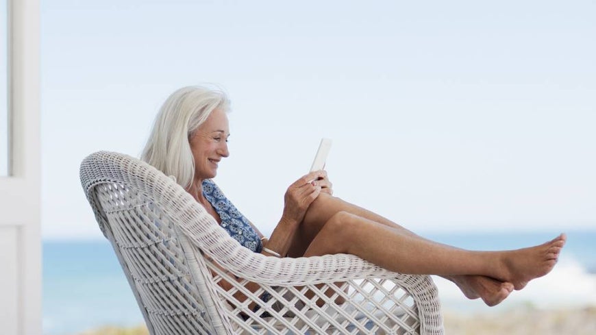 Senior woman relaxes with an ereader in a white wicker chair, near the beach. 