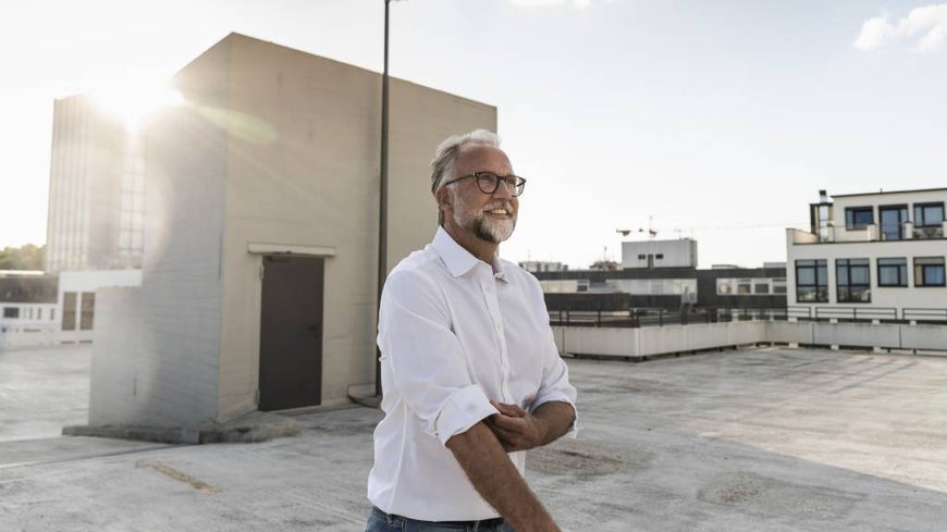 Man with white hair, beard and glasses rolling up his sleeves on a rooftop. 