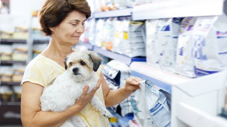 Senior woman browses pet nutrition aisle with small dog in her arms. 