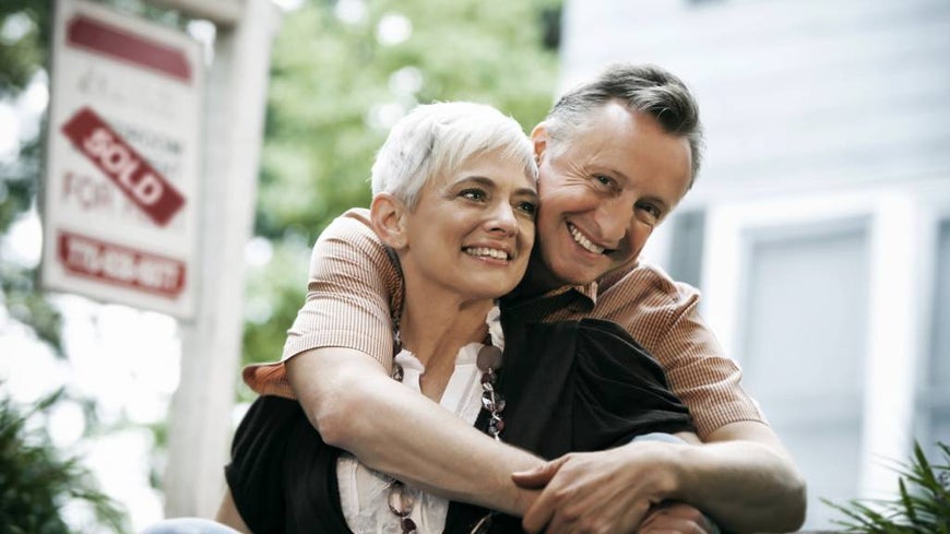 Senior couple in front of for sale sign and house. 