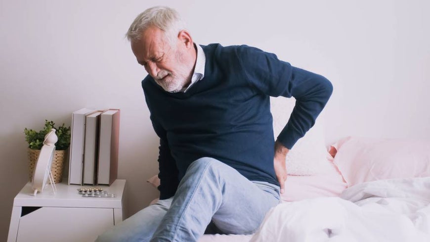 An older man sitting on the bed and holding his hip in pain