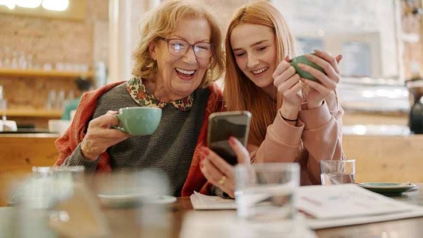 Grandmother and teenage granddaughter laugh together while looking at a smartphone.