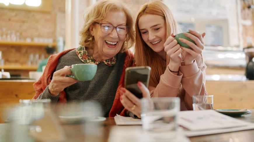 Grandmother and teenage granddaughter laugh together while looking at a smartphone. 