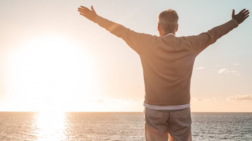Senior man on beach with outstretched hands looking to ocean.