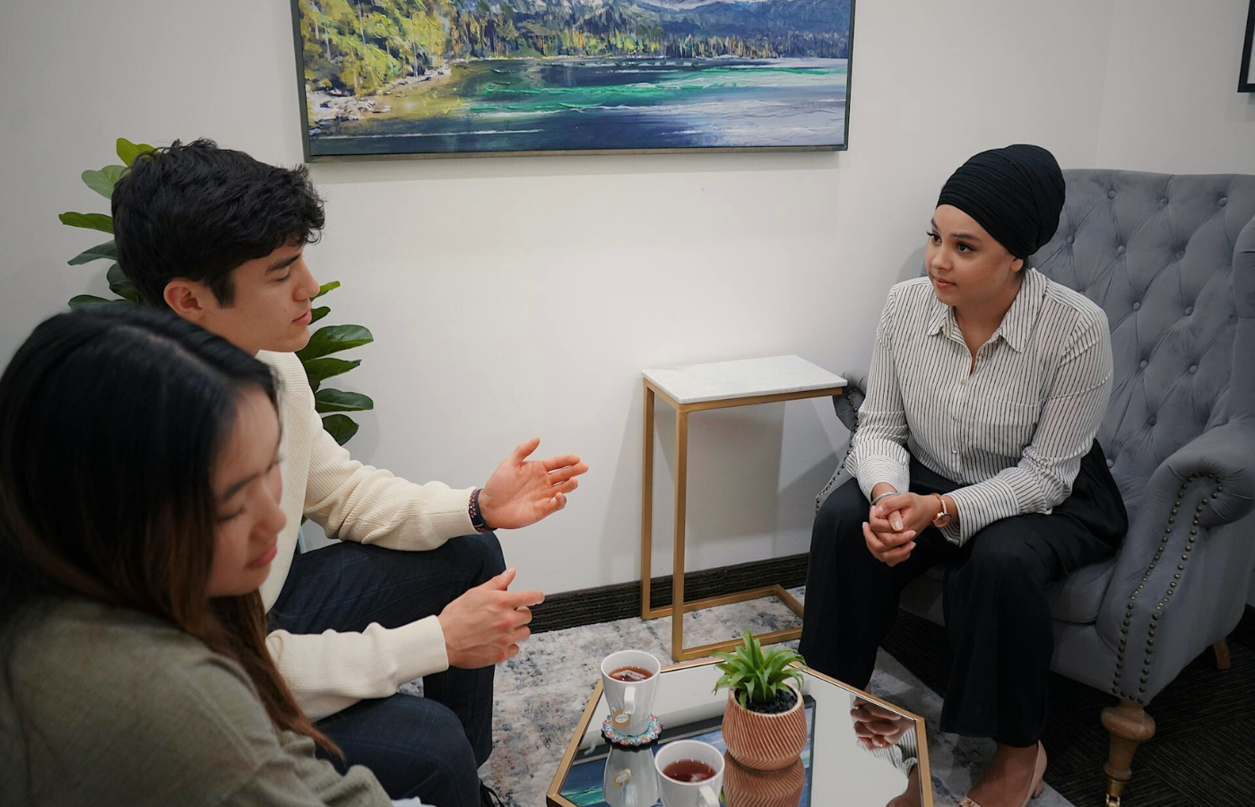 A medium shot of a couple sitting together in a warm, softly lit room. They are engaged in couples counselling session with a Psychologist - who sits opposite them. The couple appears attentive and emotionally invested, reflecting the supportive and communicative environment of this therapy session.