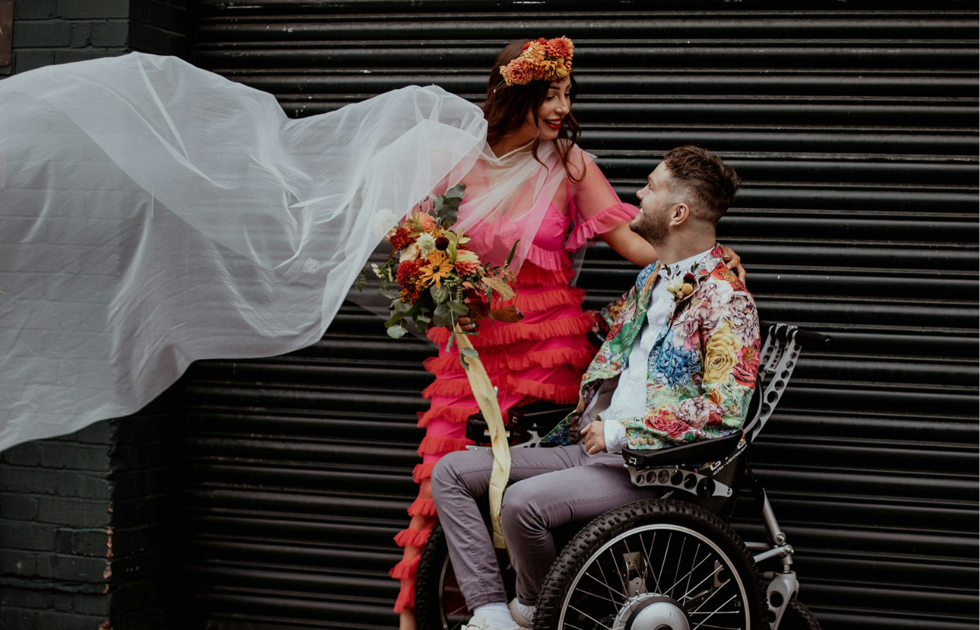 A long shot of a couple on their wedding day. They are looking into each other's eyes, and smiling. The woman is standing, while the man is seated in a wheelchair. The man wears a colourful jacket, and the woman wears a pink ombre dress and a flower crown. She is also holding a bouquet of flowers and her veil is caught by the breeze.