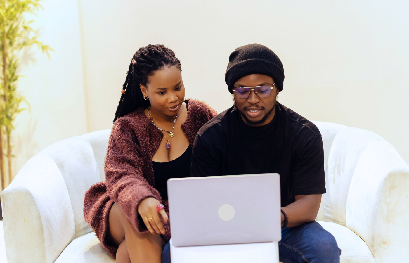 A medium shot of a couple, sitting closely together on a white love seat. They are both smiling, and and looking at the screen of a silver laptop - they are in a telehealth couples therapy session.