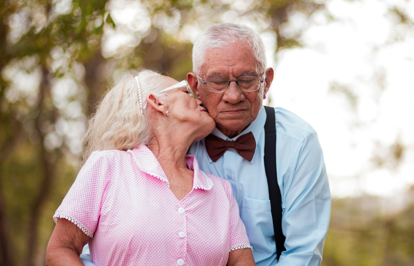 A mid-shot of a couple outdoors. The woman is encircled in the man's arms, and she leans back to kiss him on the cheek.