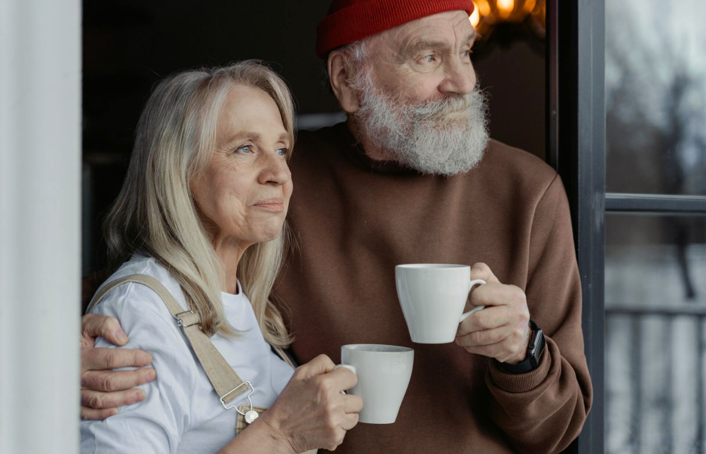 A medium shot of a couple. They gaze into the distance while they reflect on their recent couples counselling session. The man is holding his wife in a side hug, and they each hold a mug. They are wearing warm clothes and a reflection on the nearby window shows leafless trees and snow-covered ground.