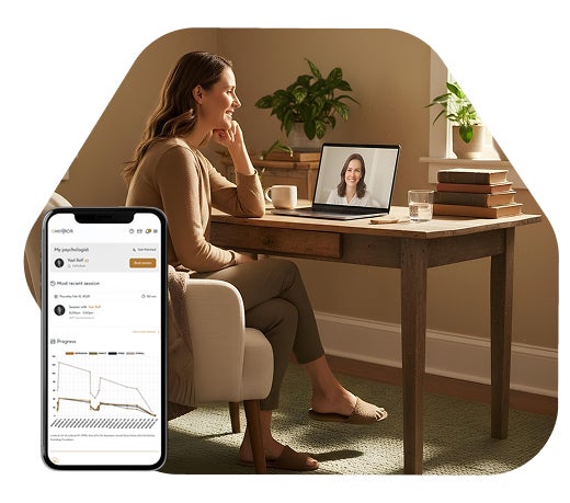 A woman smiles while having an online video session at a wooden desk with a laptop, books, and a glass of water, with a smartphone displaying a My Mirrors online telehealth platform