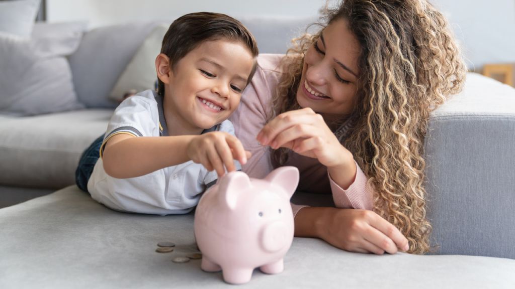 happy mother and son placing money in a piggy bank while laying on a couch