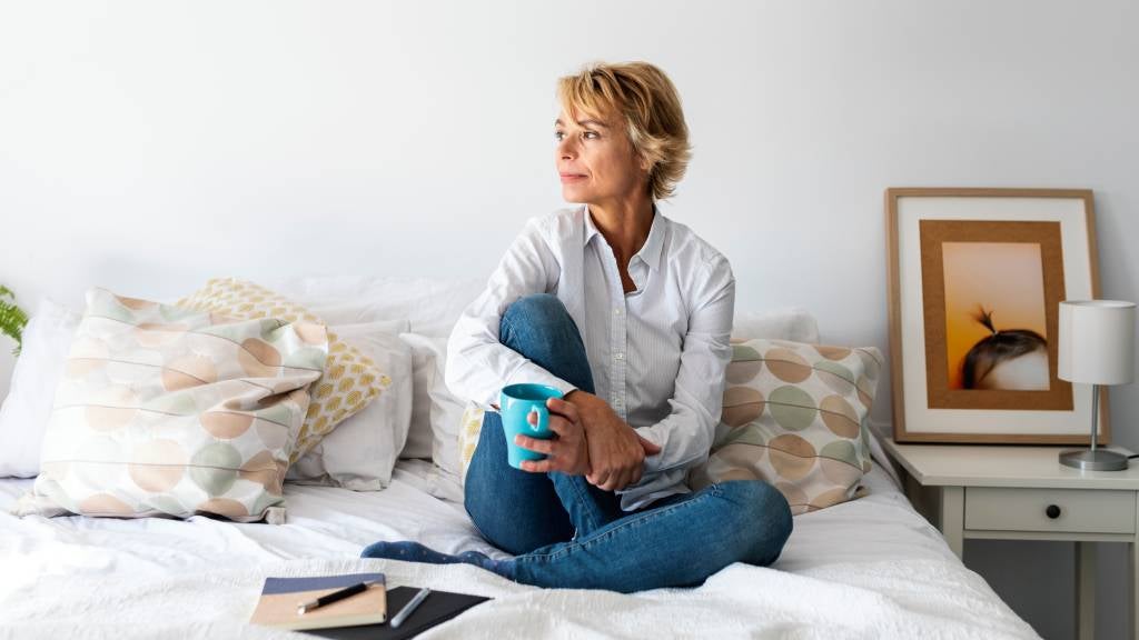 Mature woman sits on her bed with cup of tea and notepad and pens.
