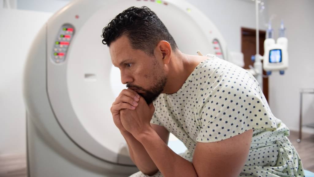 A worried looking man sits clasping his hands in front of a CT scanner