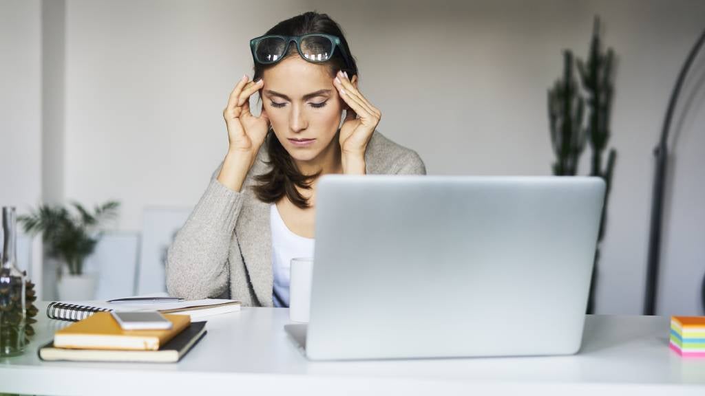 A tense woman with eyes closed sits at her desk and presses her temples